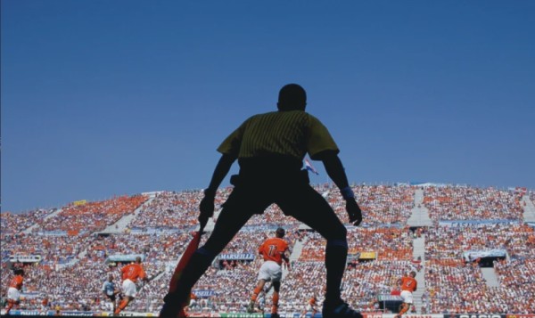 Photograph of the unlit back of a man holding a flag on a field, watching well lit players running on a field, looking up at a large crowd of people in the stands and a blue sky.