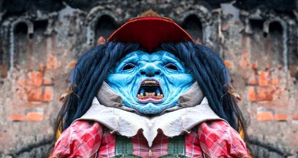 A sculpture of a distressed blue monster wearing farm clothing and a wig, in front of arched stone structures.