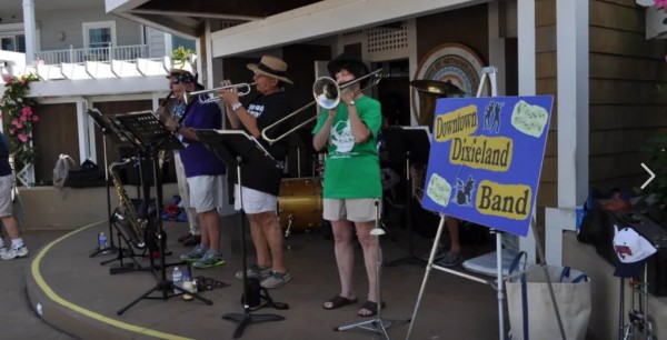 Four people on a platform in front of a building playing instruments, next to a blue sign stating 'Downtown Dixieland Band.'
