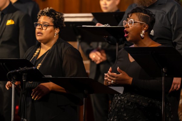 A black person with glasses and short hair in front of a music stand, singing on stage.