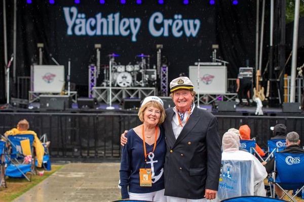 Twp people smiling, standing in front of a stage with band equipment and a sign stating 'Yachtley Crew.'