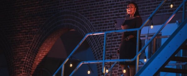 A black woman singing on a metal staircase in front of a brick wall with an archway.