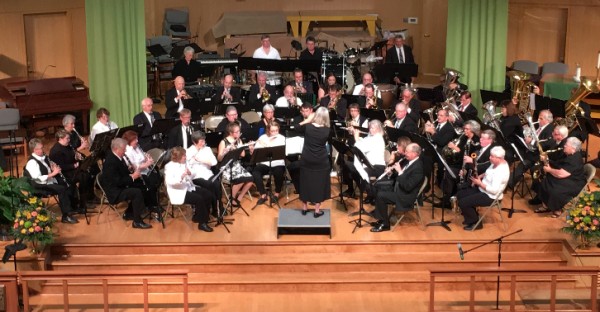 A concert band on stage wearing black and white, playing various instruments and being conducted by someone with long blonde hair and a black dress.