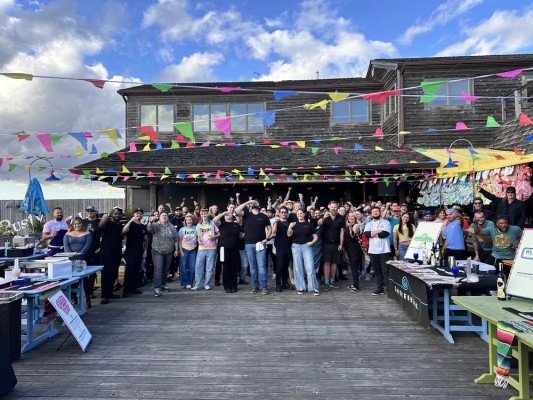 A group of roughly 100 people on the deck of a wood building, celebrating, with merchant tables surrounding them.