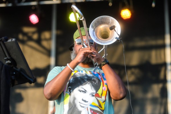 A black man with a gray beard, playing the trombone on an outdoor stage. He is wearing a colorful Aretha Franklin t-shirt.
