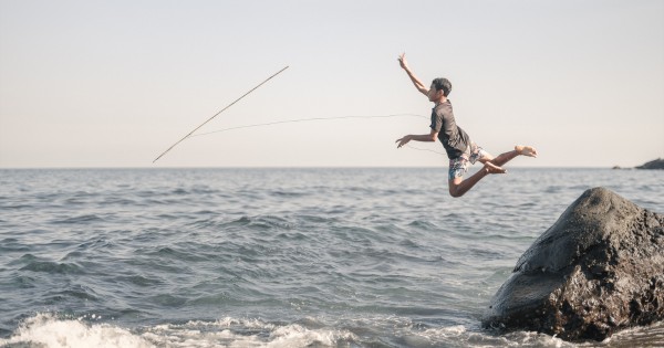 A boy jumping off of a rock and throwing a fishing rod  into the ocean.