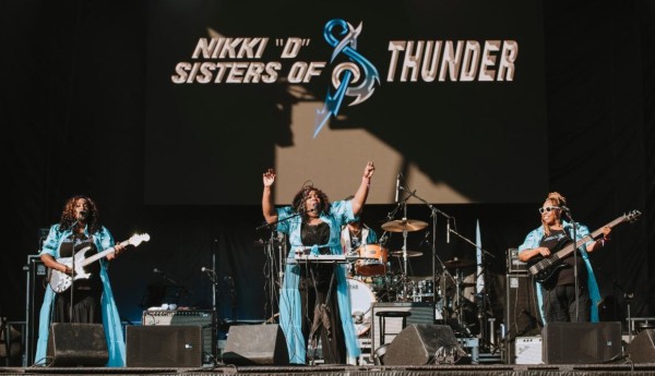 Four women on stage performing music, wearing long black dresses and long light blue coats over them.