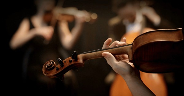 A hand playing a violin with other blurred musicians in the background.
