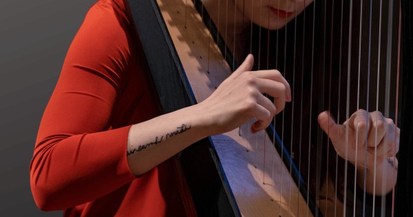 A woman in a red shirt with a tattoo on her arm playing the harp, in front of a black background.