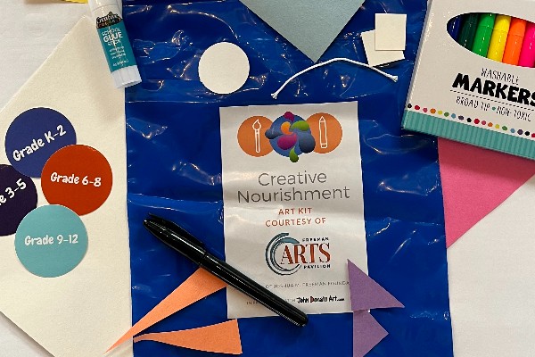 A blue plastic bag with a label reading "Creative Nourishment Art Kit Courtesy of Freeman Arts Pavillion" surrounded with various art supplies including cut bits of colored paper, markers, a pen, and a glue stick. There are stickers beside the bag that read: Grade k-2, Grade 6-8, Grade 9-12, and Grade 3-5.